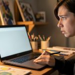 An artist at a desk with a tablet and stylus looking at a laptop screen with a concerned expression