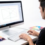 A designer at a computer preparing a print layout with crop marks and bleed on screen next to a printed proof