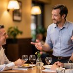A restaurant manager talking calmly with a seated guest at a table in a warmly lit dining room