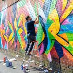 An artist on a step ladder painting a colorful abstract mural on an outdoor brick wall