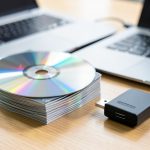 A stack of compact discs beside a laptop and a small external drive on a desk, one disc catching rainbow light