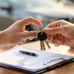 A person handing over car keys next to a clipboard with paperwork and a set of documents on a desk