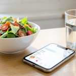 A phone showing a simple food diary app next to a bowl of healthy food on a kitchen table