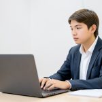 A student at a tidy desk holding a laptop webcam up to scan the room before an online exam