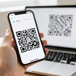 A phone held up to scan a QR code displayed on a laptop screen to log in