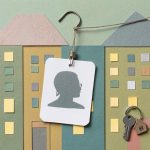 Cut paper collage of a blank ID card with a silhouette portrait hanging from a fishing hook in front of colorful paper apartment buildings, with a house key on a ring