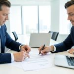 Two business colleagues reviewing a software contract on a laptop and printout at a conference table