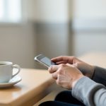 A person's hands typing on a smartphone at a table with a coffee cup in a calm hospital waiting area