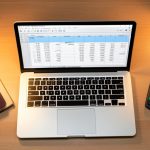 A laptop showing a budget spreadsheet next to a passport, a notebook, and a small calculator on a desk