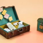 Papercraft illustration of an open vintage suitcase filling with paper envelopes, a hotel and paper airplanes, beside a locked paper mailbox